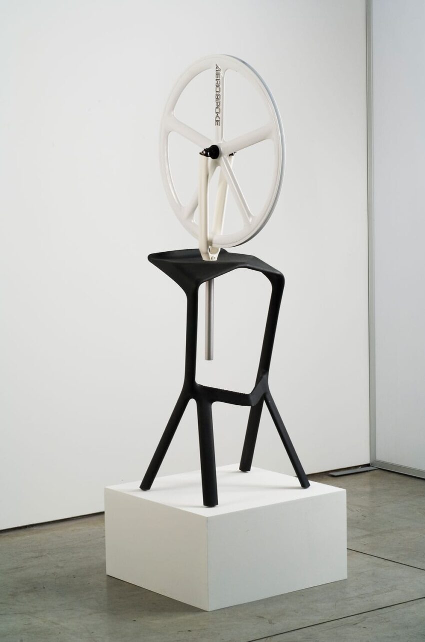 Modern black and white sculptural chair with a large circular wheel on top displayed on a white pedestal.