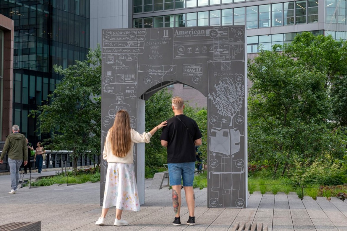 Two people stand in front of a large artistic archway with drawings and text, set outdoors in a modern urban area.