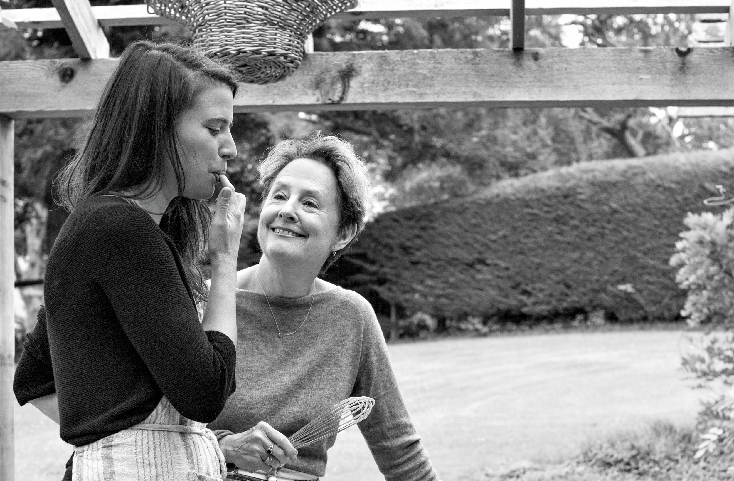 Two women smiling outdoors, one tasting from a spoon while the other holds a whisk, under a wooden pergola in a garden setting.