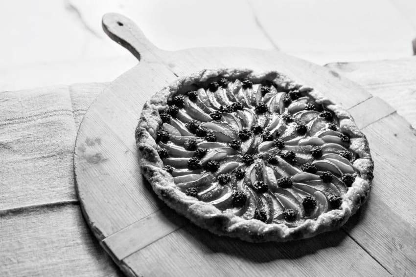 Black and white image of a fruit tart on a wooden serving board with a decorative crust and neatly arranged toppings.