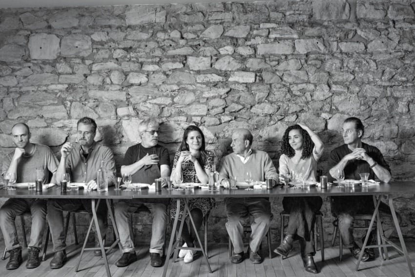 Group of seven people sitting at a long table against a stone wall.