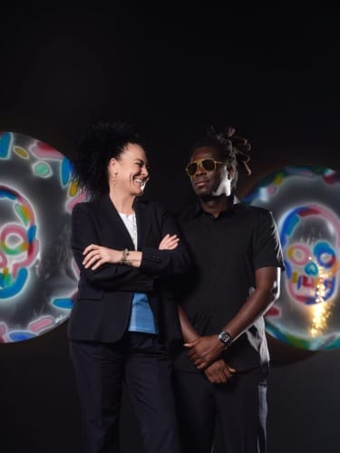 Two people smiling in front of colorful skull artwork on a dark background.