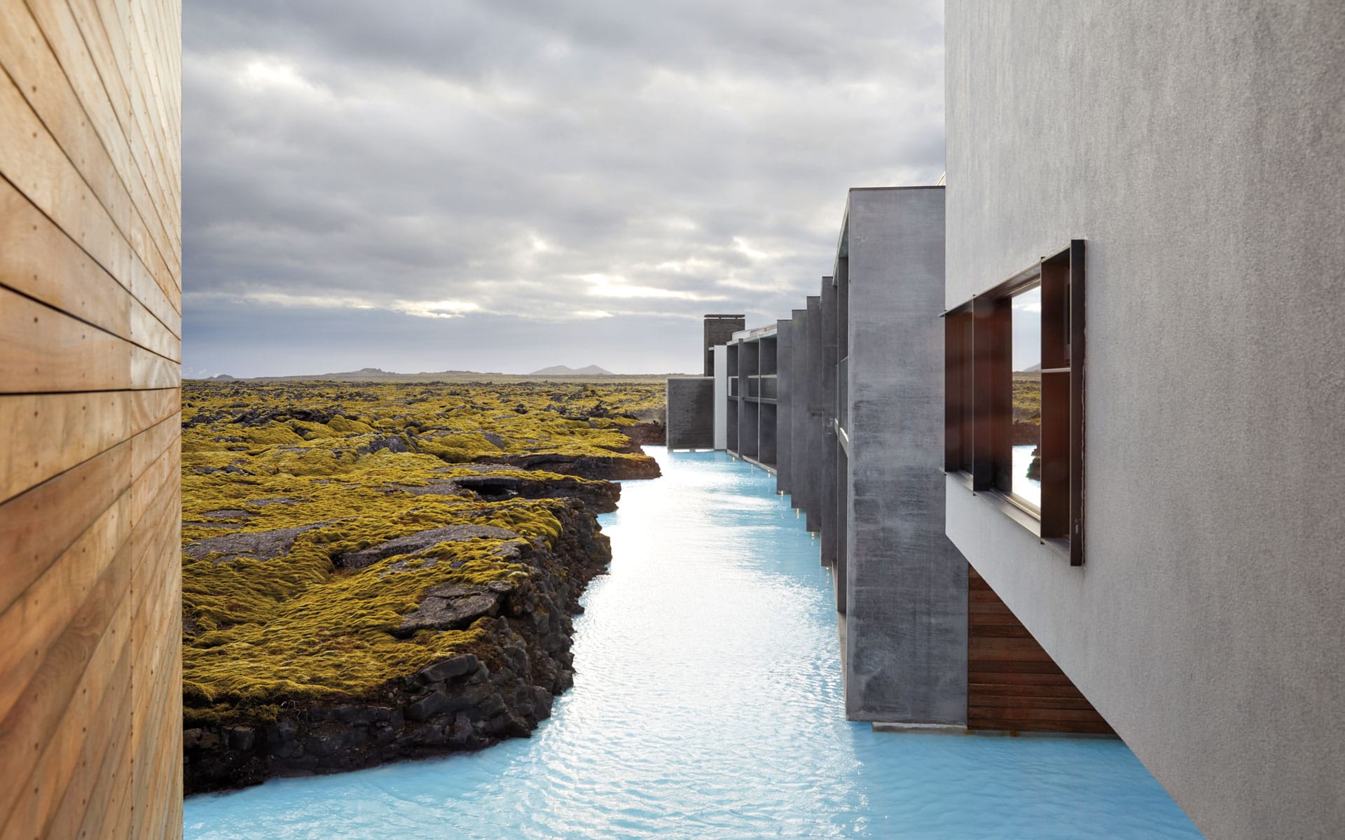 Modern building overlooking blue geothermal water surrounded by lava rocks and moss under a cloudy sky.