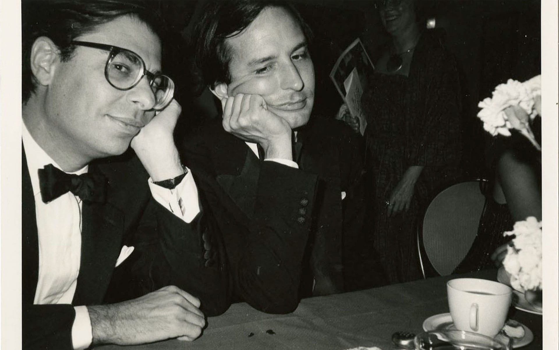 Two men in formal attire sitting at a table, resting their heads on their hands, with a coffee cup and flowers nearby.