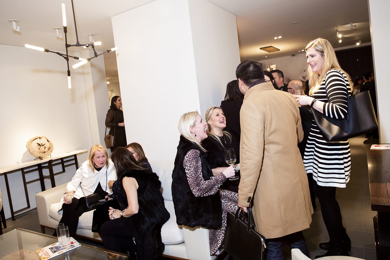 Group of people socializing in a modern living room setting, some seated and others standing, engaged in conversation and laughter.