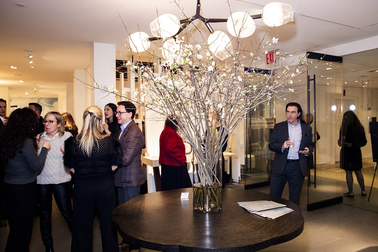 Elegant indoor gathering with people socializing around a large table centerpiece of blooming branches in a modern setting.
