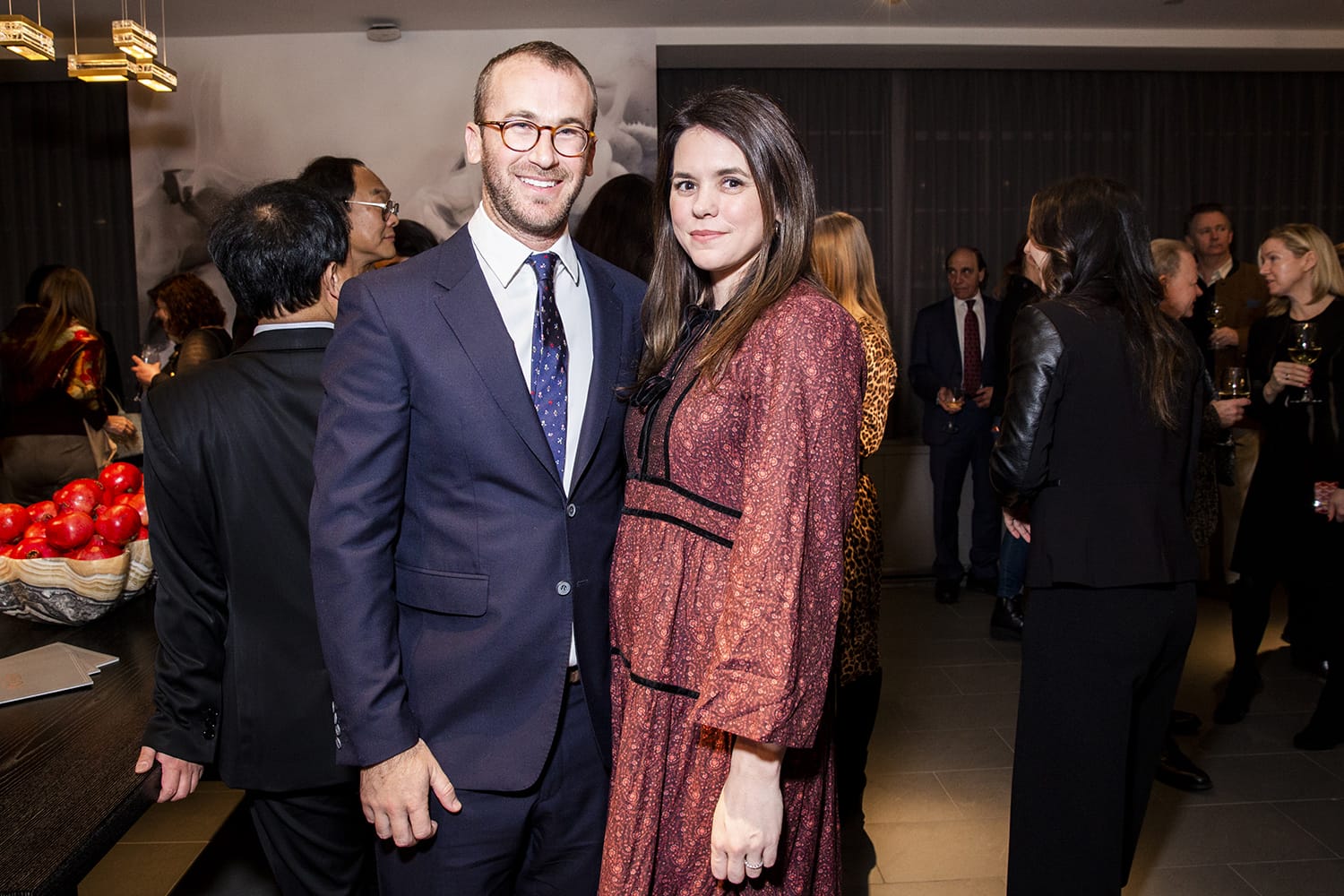 A man and woman smiling and posing at a social gathering with other people in the background.