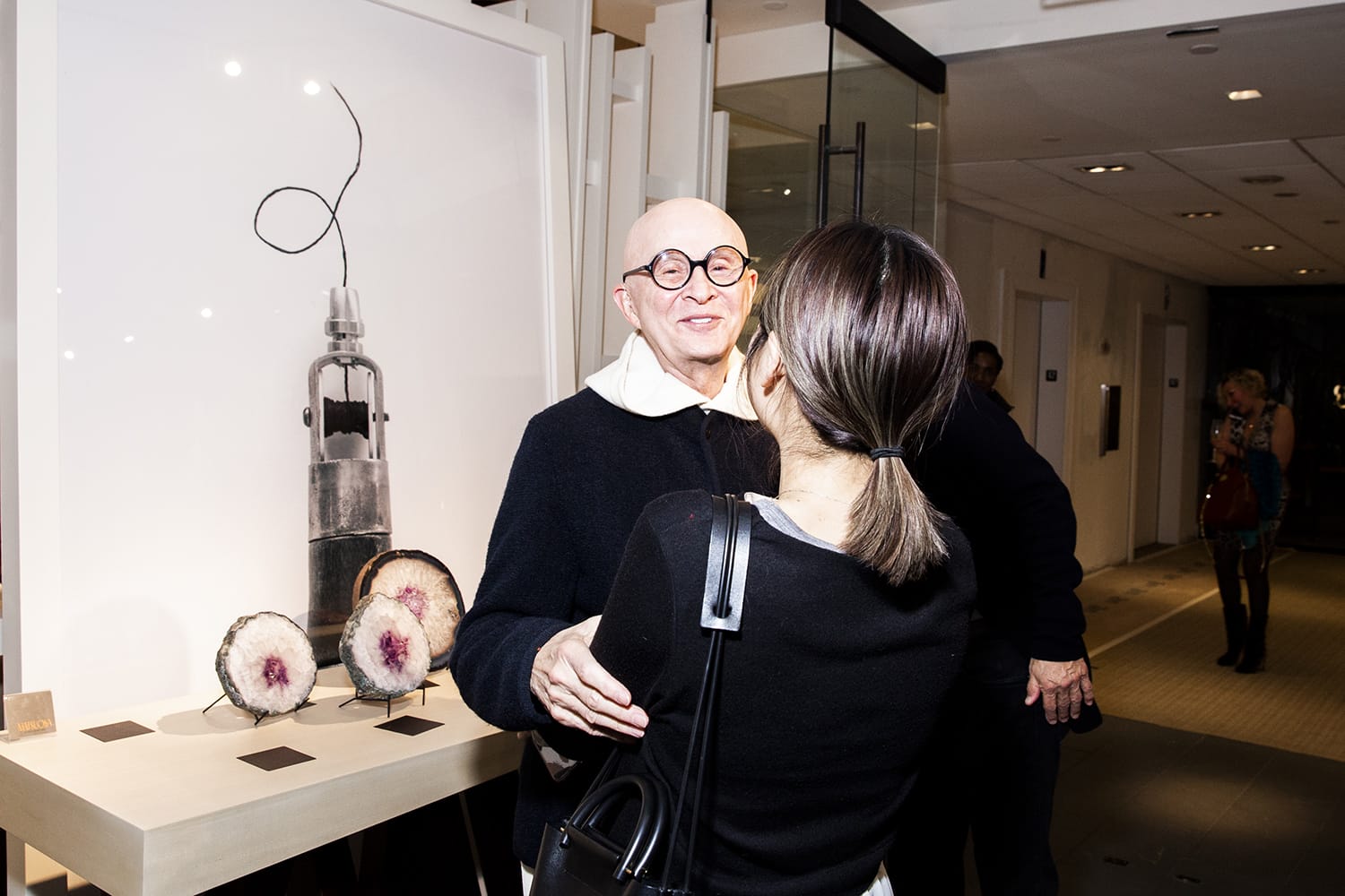 A person with glasses smiles and interacts with another person in an art exhibit with sculptures in the background.