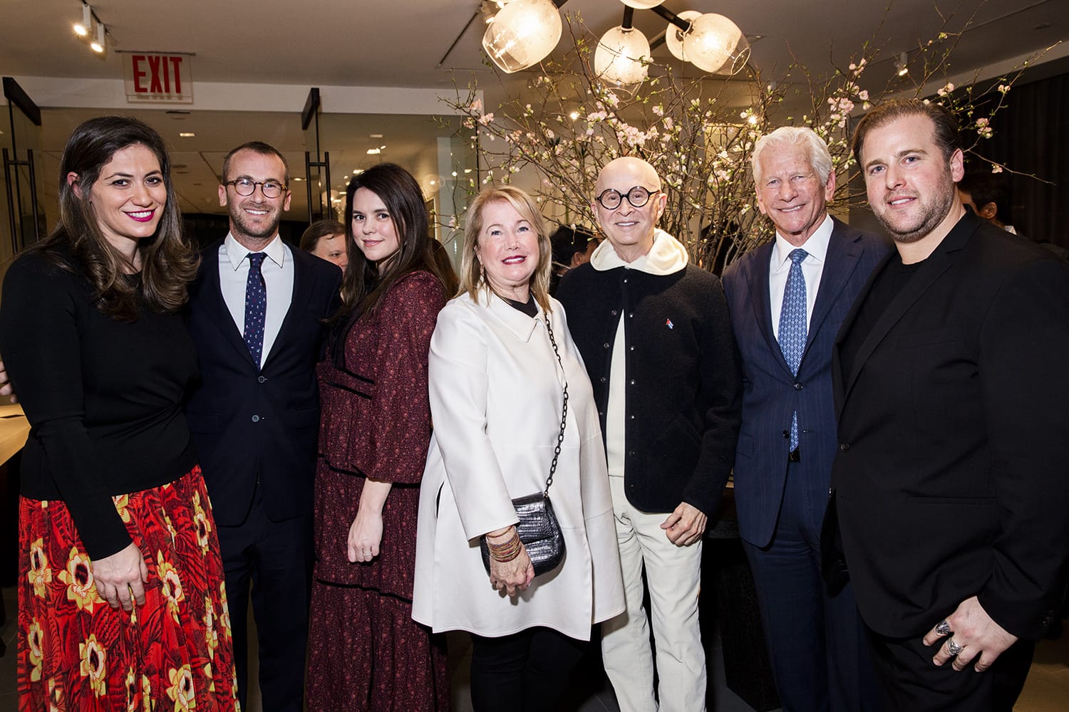 Group of elegantly dressed people posing together at a formal event with floral arrangement in the background.