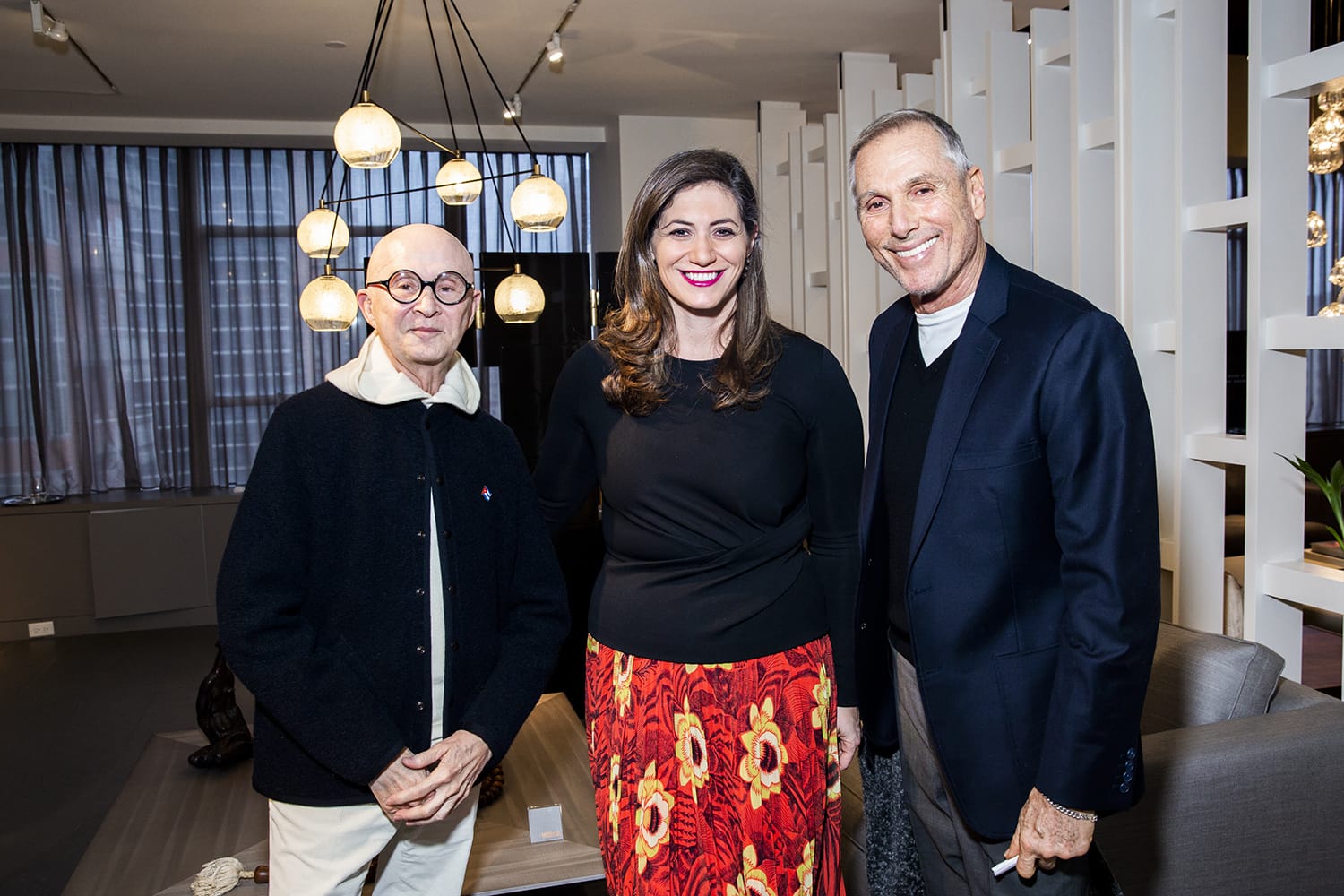 Three people smiling, standing together in a modern room with hanging lights and a patterned red skirt worn by the person in the center.