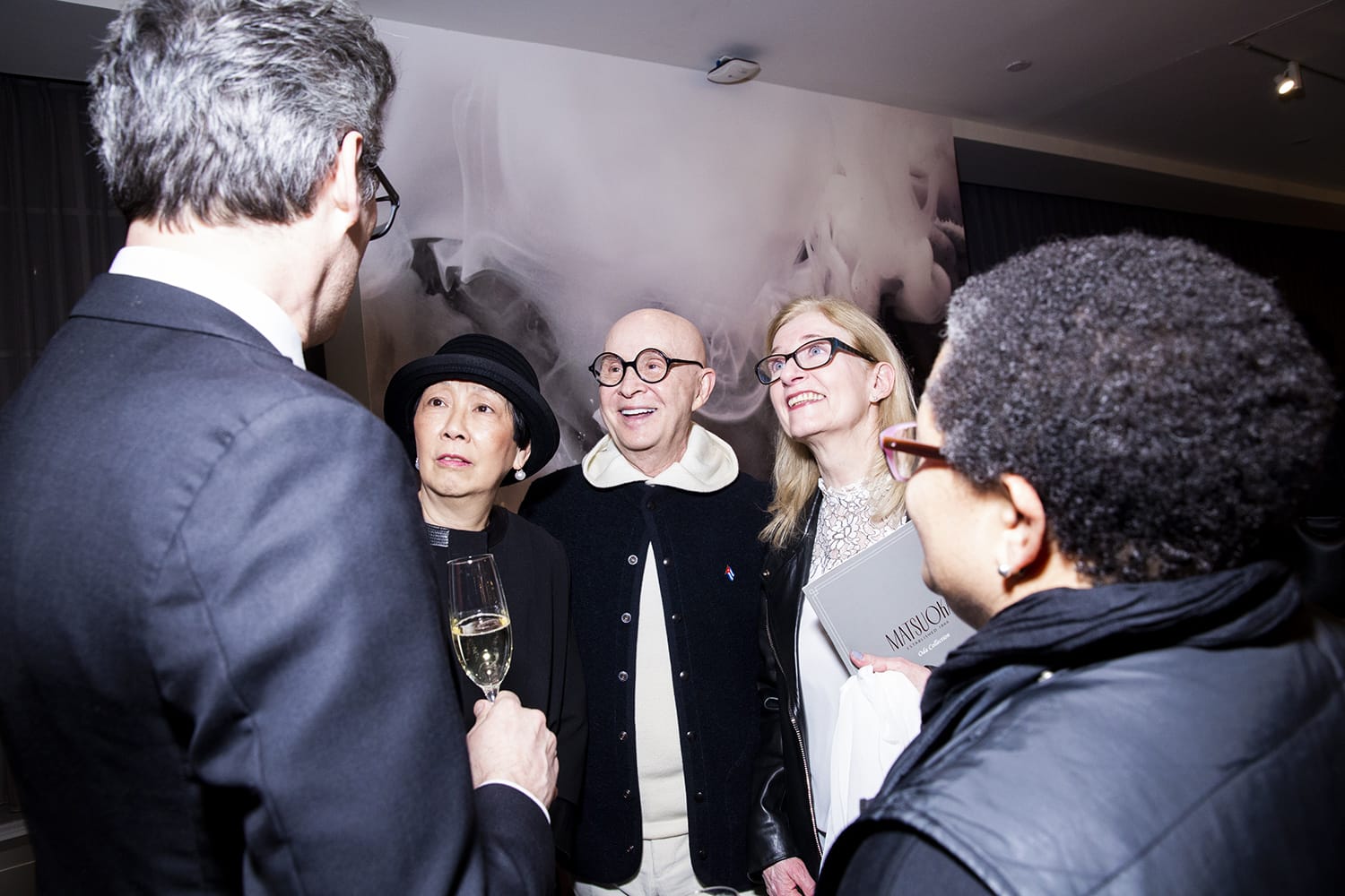 Group of people smiling and talking at a social gathering, one holding a glass of champagne, with an abstract art backdrop.