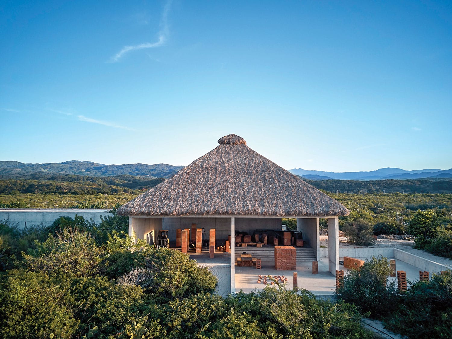 A thatched roof pavilion surrounded by lush greenery with mountains in the background under a clear blue sky.