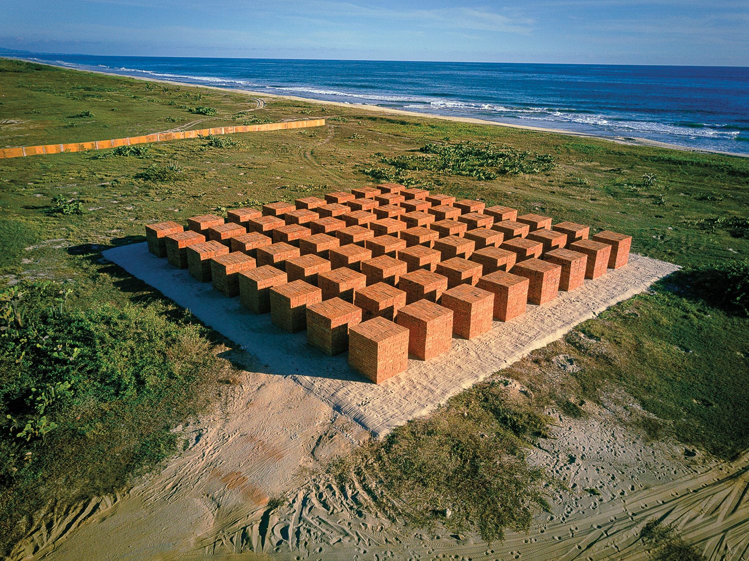 Outdoor art installation featuring a grid of brick cubes near a coastline, surrounded by grass and sandy terrain.