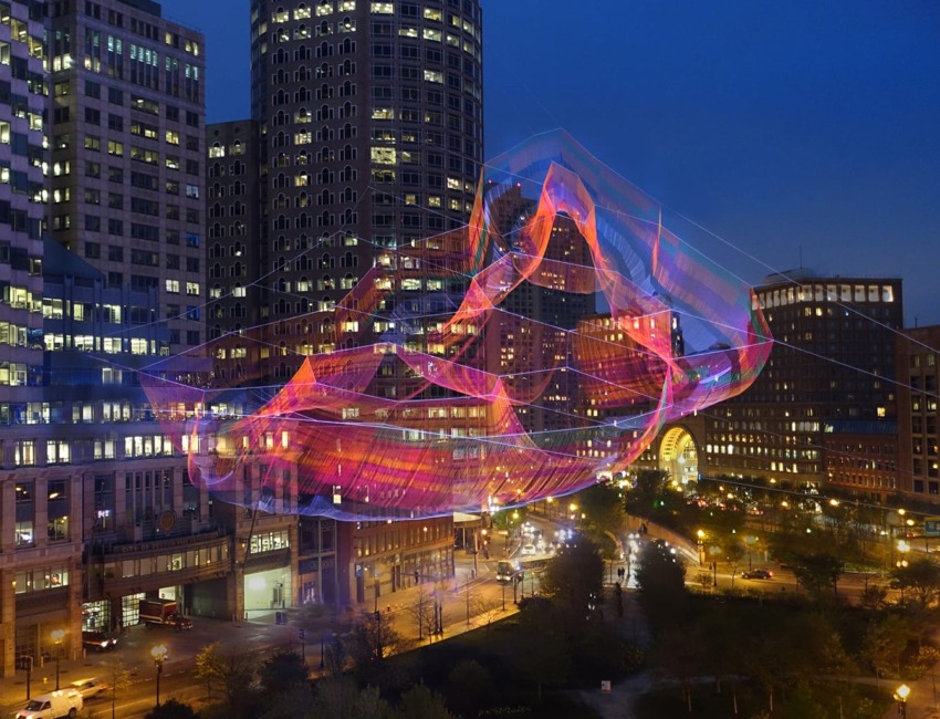 Colorful illuminated art installation above a city plaza at night, with tall buildings in the background.