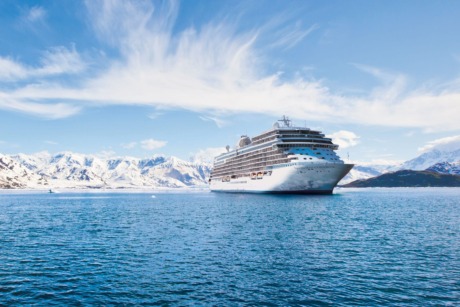 Cruise ship sailing on calm blue waters with snow-capped mountains in the background under a clear blue sky.