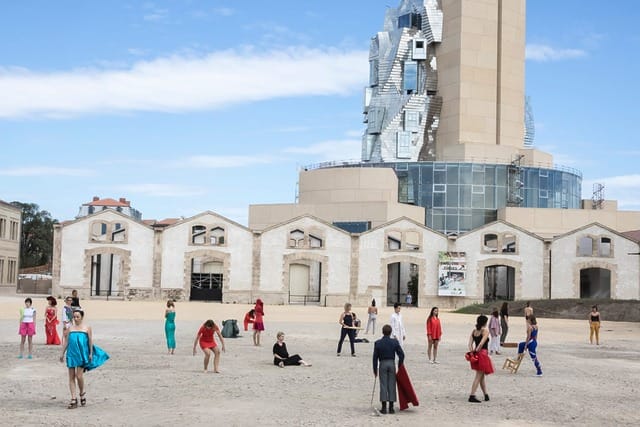 People in colorful outfits stand on an open space in front of modern and historic architecture under a blue sky.