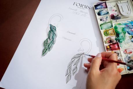 Hand sketching elegant leaf-shaped earrings with a watercolor palette nearby.