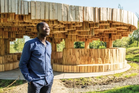 Man standing in front of a unique wooden structure with interlocking beams, surrounded by greenery on a sunny day.