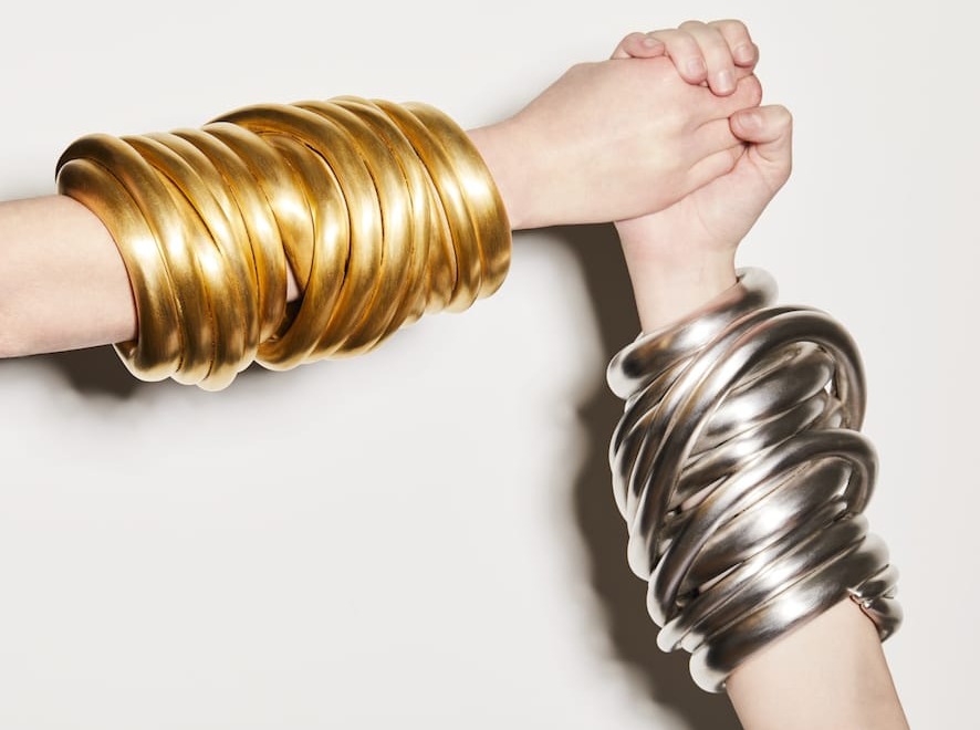 Two hands with stacked metallic bangles, one gold and one silver, holding each other against a white background.