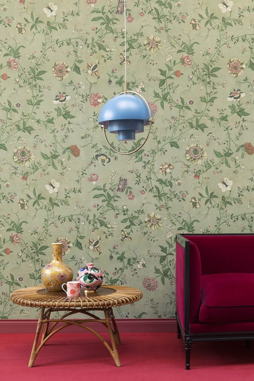 Vintage floral wallpaper, blue pendant lamp, wicker table with colorful vases, and a red couch on a red carpet.