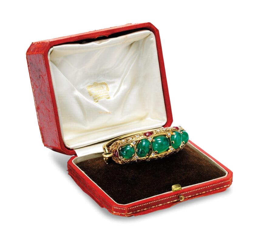 Vintage gold bracelet with green stones and a red gem, displayed in an open red velvet and satin jewelry box.