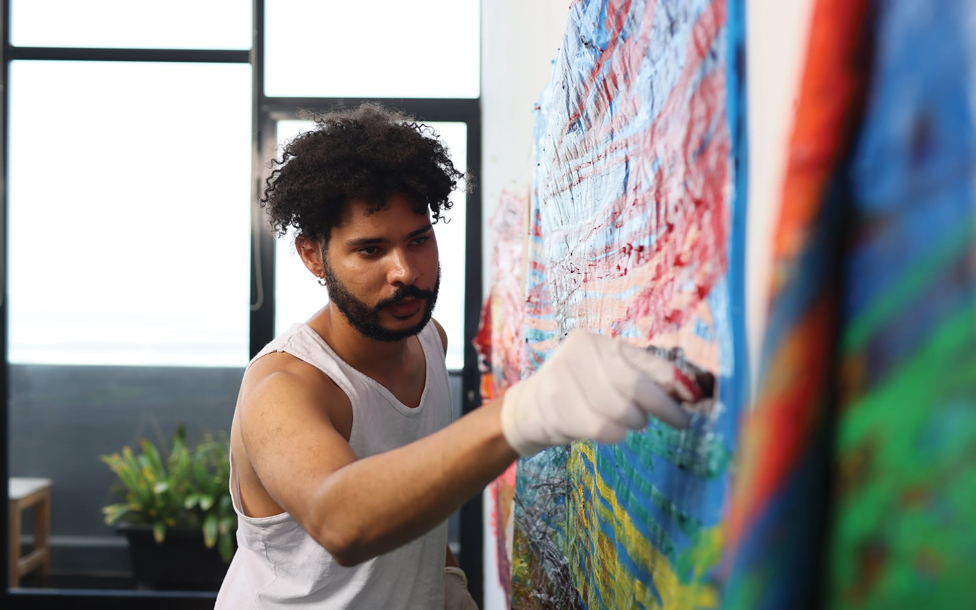 Man painting a vibrant abstract mural indoors with a focused expression, wearing a white tank top and gloves.