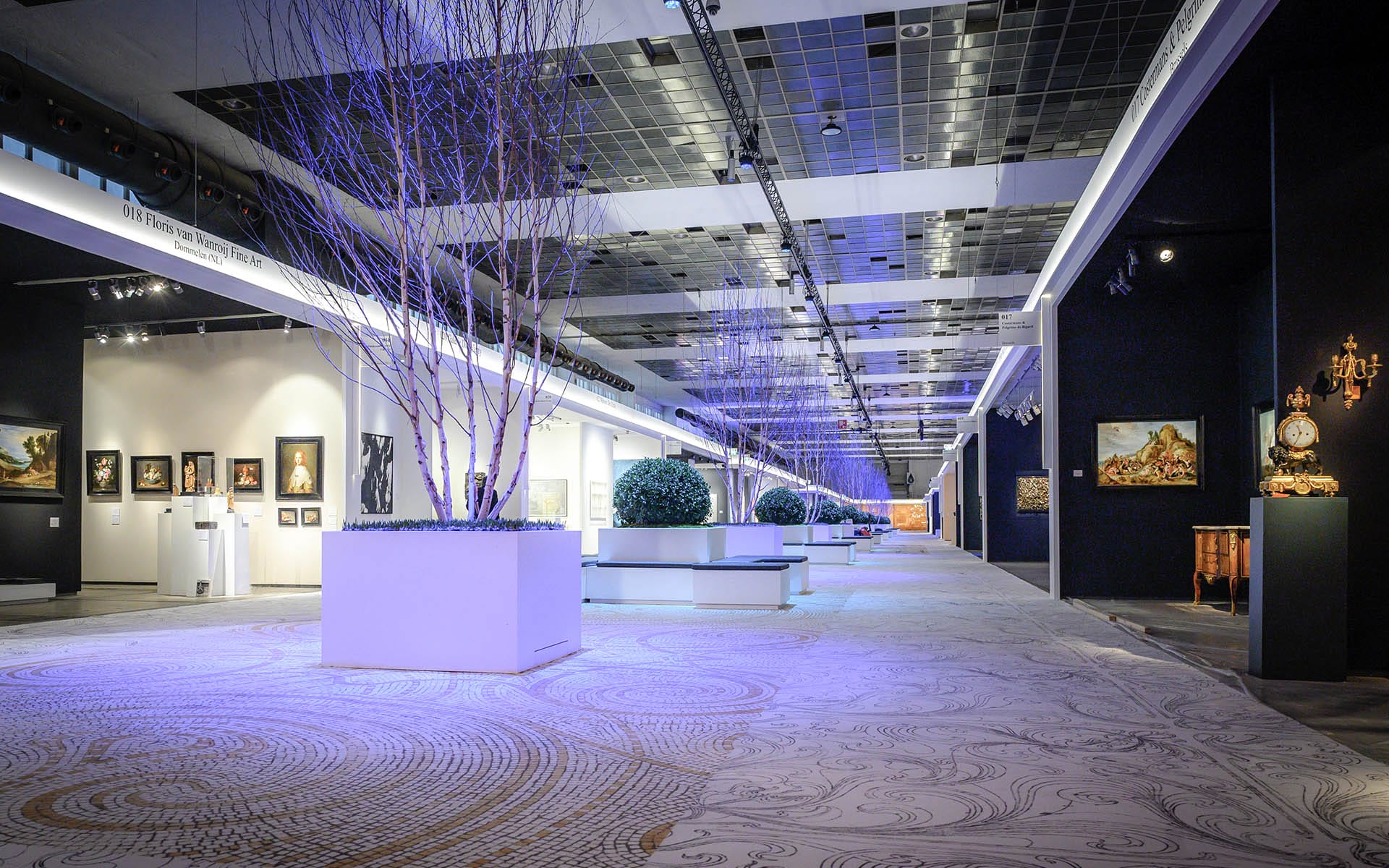 Spacious modern art gallery with illuminated installations, tree displays, and paintings on walls under blue ambient lighting.