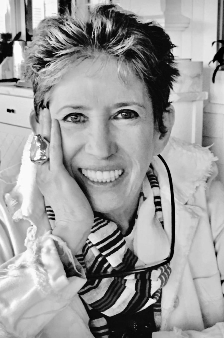 Woman smiling warmly, resting chin on hand, wearing a striped scarf and large ring with short hair in a cozy setting.