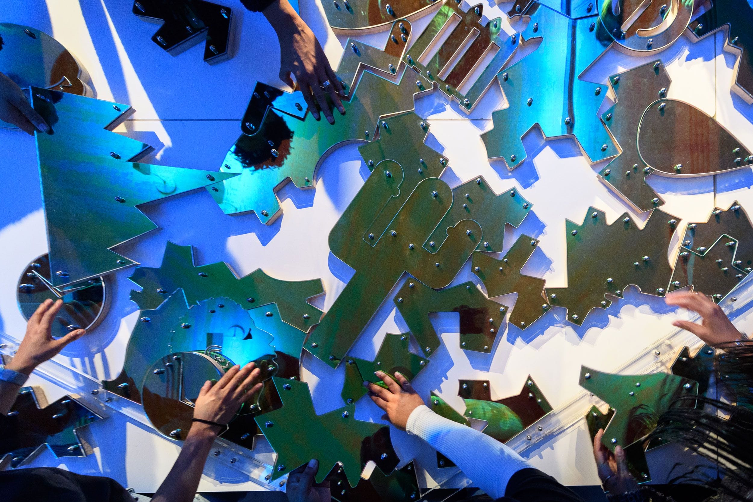 Hands assembling colorful metal puzzle pieces with various shapes on a blue-lit wall.