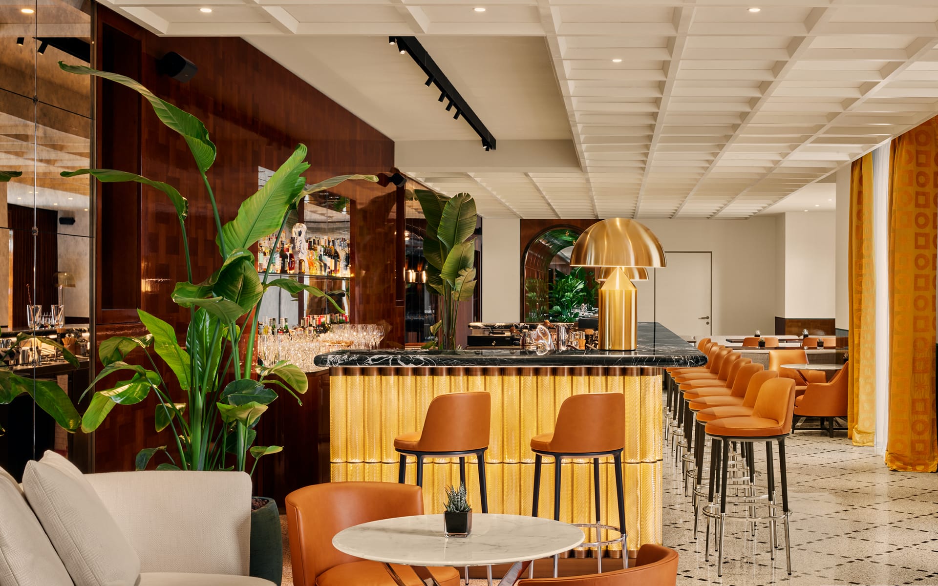 Modern bar interior with orange chairs, marble tables, lush green plants, and ambient lighting.
