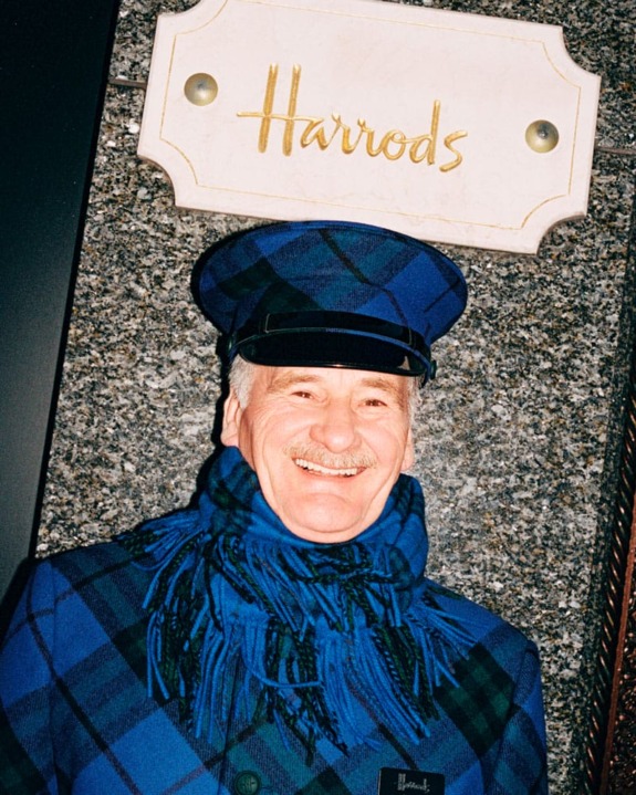 Man in blue plaid uniform smiling in front of Harrods sign.