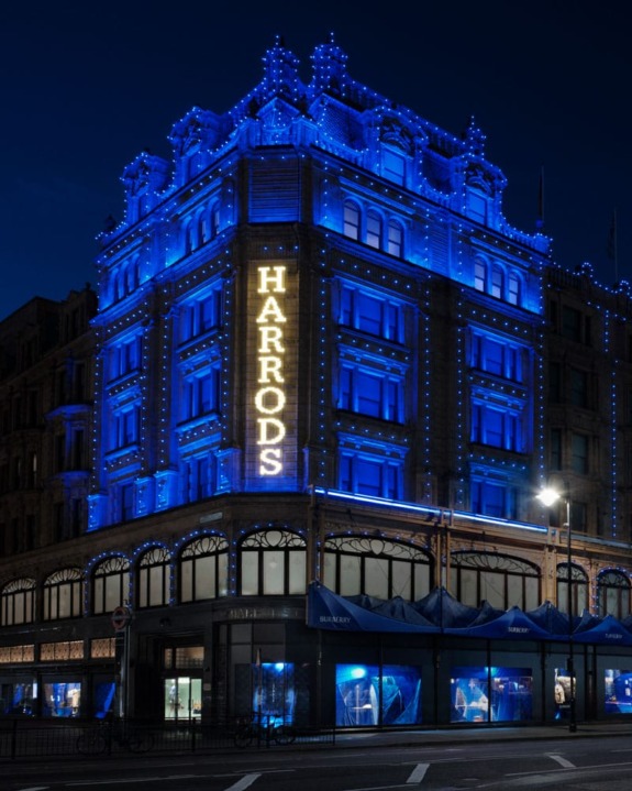 Harrods department store exterior at night illuminated in blue with glowing signage on a street corner.