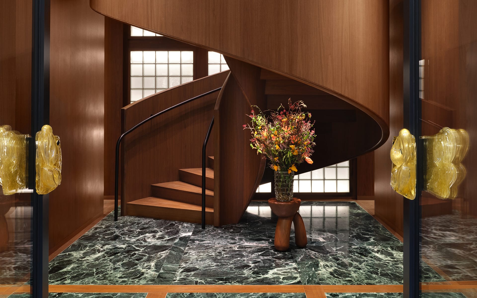 Spiral wooden staircase with a vase of colorful flowers on a marble floor, framed by two glass doors with gold handles.