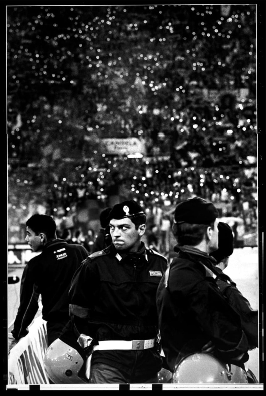 Police officers in uniform stand on a dimly lit stadium field with a crowd in the background.