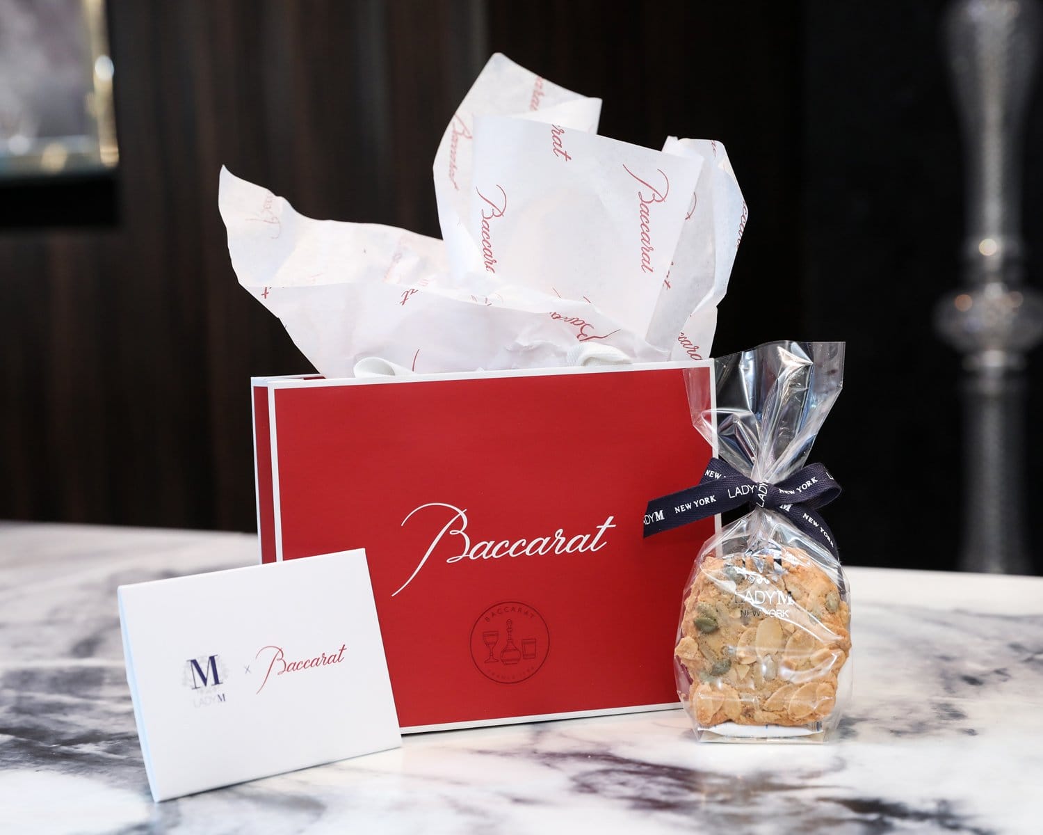 Red Baccarat gift bag with tissue paper, packaged cookies in clear bag, and a white card on a marble table.