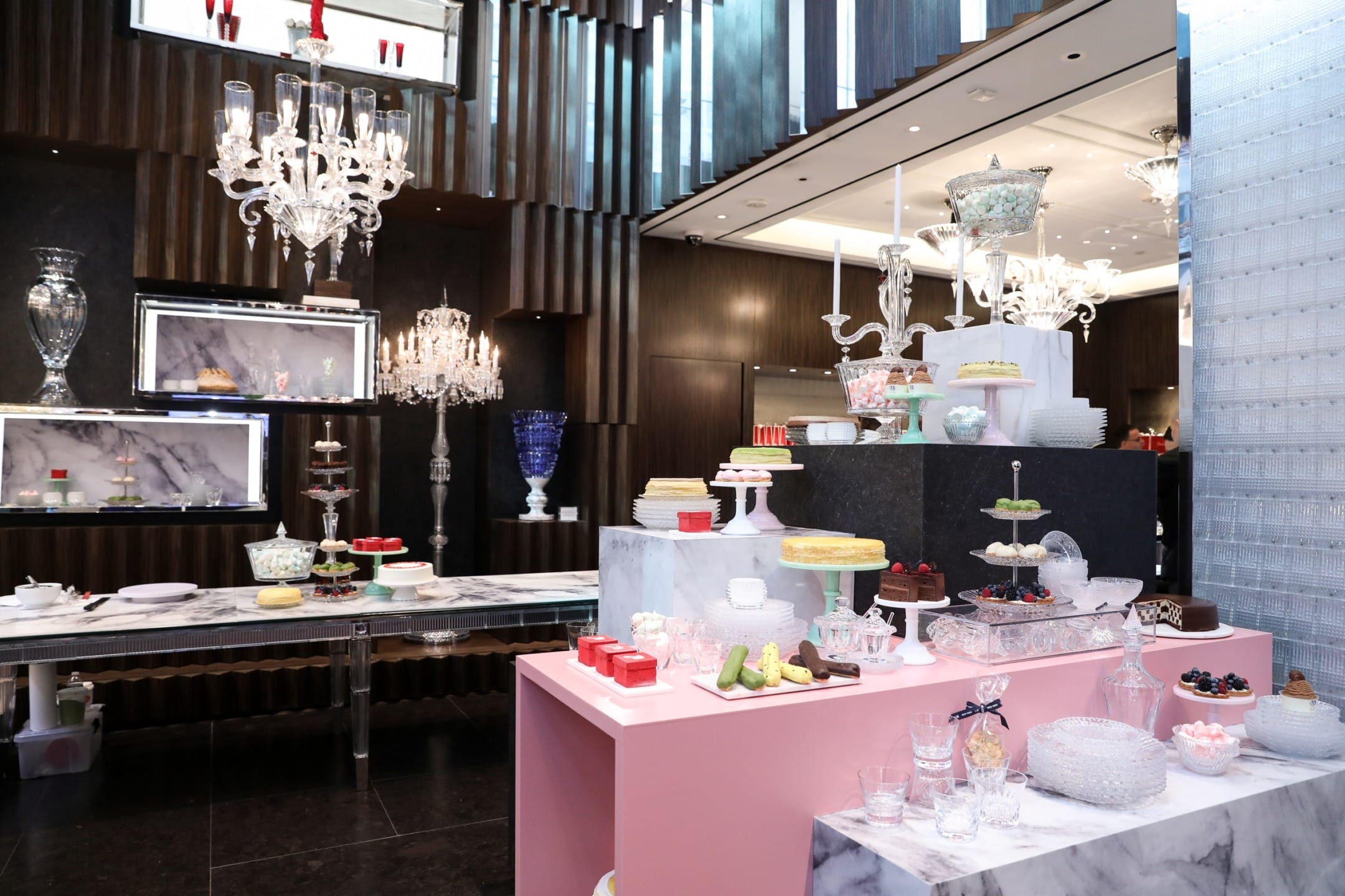Elegant dessert display with cakes, pastries, and glassware in a luxurious setting with chandeliers and modern decor.