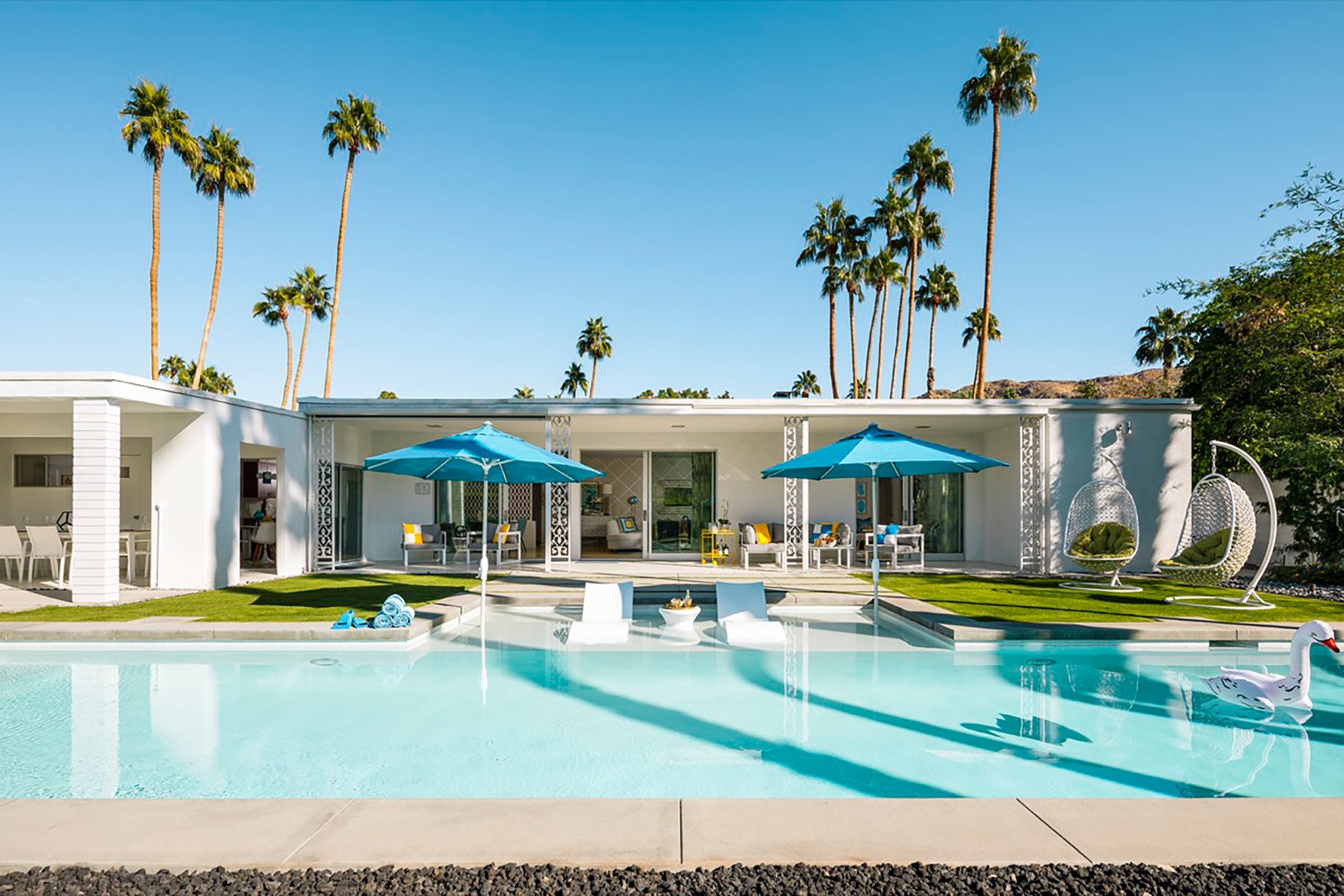 Modern house with large pool, blue umbrellas, and palm trees under a clear sky