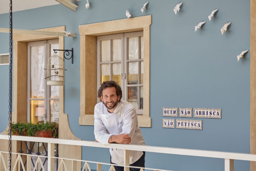 Chef in a white coat stands on a balcony with blue walls, decorative birds, and a Portuguese quote on tiles.