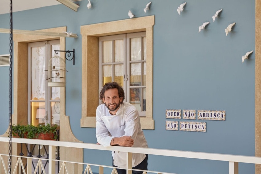 Chef in a white coat stands on a balcony with blue walls, decorative birds, and a Portuguese quote on tiles.