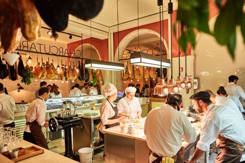 Chefs and staff working in a busy charcuterie kitchen with hanging meats and bright lighting.