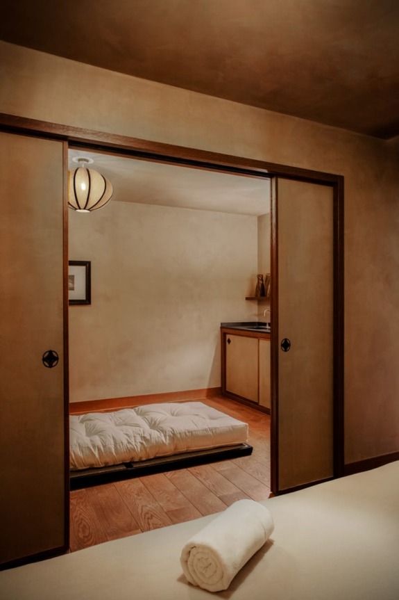 Japanese-style room with futon on wooden floor, soft ambient lighting, rolled towel in foreground, and a small kitchenette.