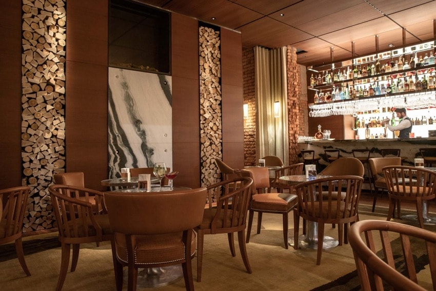 Cozy restaurant interior with wooden decor, round tables, and a well-stocked bar in the background.