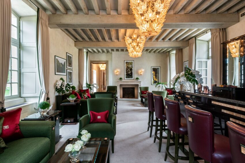 Elegant living room with green and burgundy furniture, chandeliers, large windows, and a fireplace at the center.