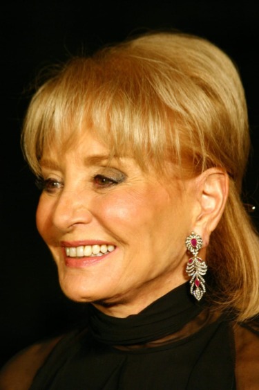 A woman with blonde hair wearing elegant jewelry and a black top, smiling against a dark background.