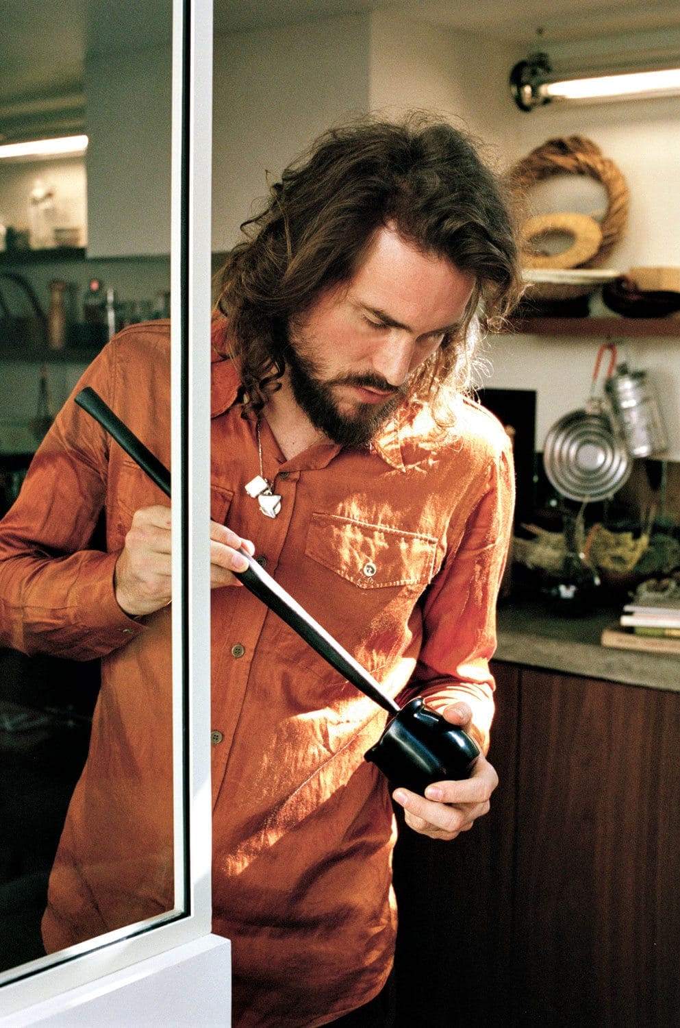 Man in an orange shirt examines a black object in a kitchen setting, with various items visible in the background.