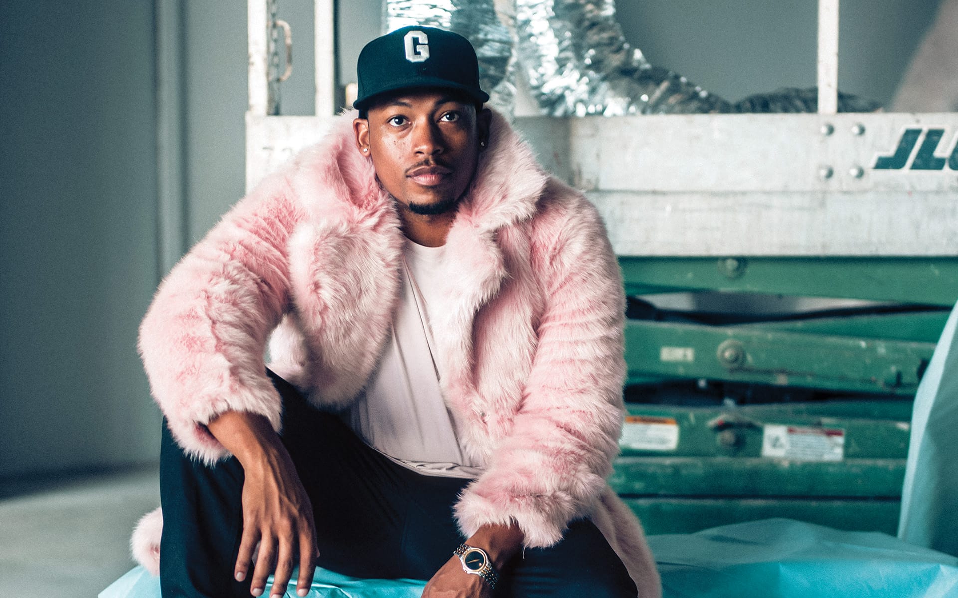 Person in a pink fur coat and black cap sitting on a construction lift indoors.