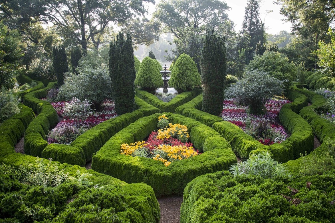 Intricately designed garden with vibrant flowers, manicured hedges, and a central fountain on a sunny day.