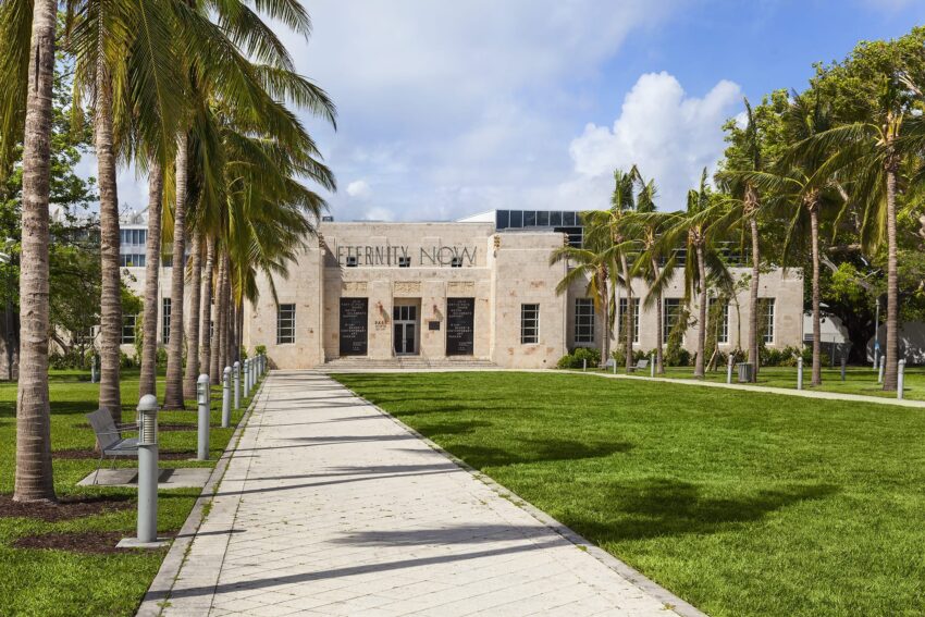 Art Deco building with "Eternity Now" sign, palm trees lining a pathway, bright green lawn, and partly cloudy blue sky.