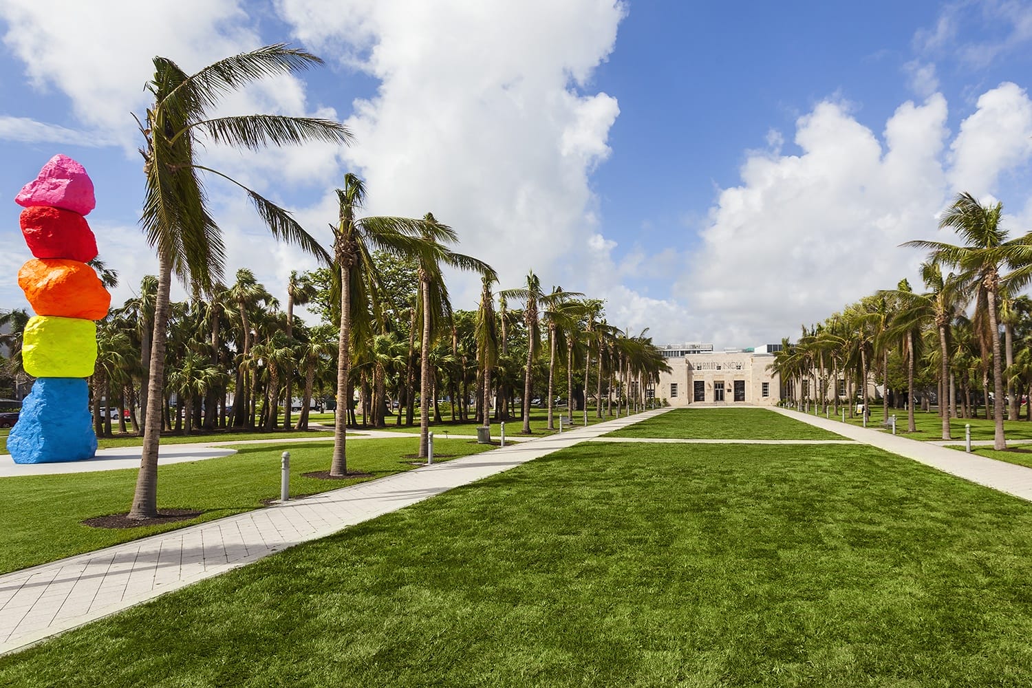 Colorful stacked rock sculpture and palm-lined pathway in a park setting under a bright blue sky.