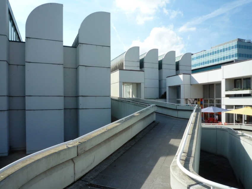 Modern architecture of a building complex with curved roof elements and a pedestrian walkway under a clear blue sky.